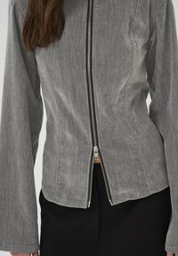 Grå, tekstureret jakke med en central lynlås med sort kant, brede ærmer og en kortet nederkant. Fremstillet af et blødt, vævet materiale.