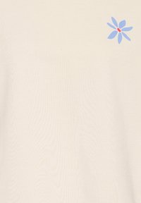 T-shirt beige clair en tissu doux présentant un motif de fleur bleue avec un centre rouge sur le côté supérieur gauche. Texture lisse.