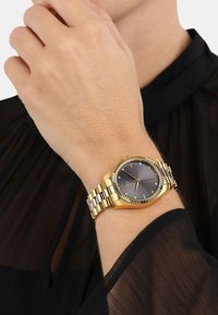 Goldene Uhr mit rundem Ziffernblatt, grauem Zifferblatt und silberfarbenen Zeigern. Verfügt über ein gemustertes Gliederarmband und dekorative Akzente am Lünette.