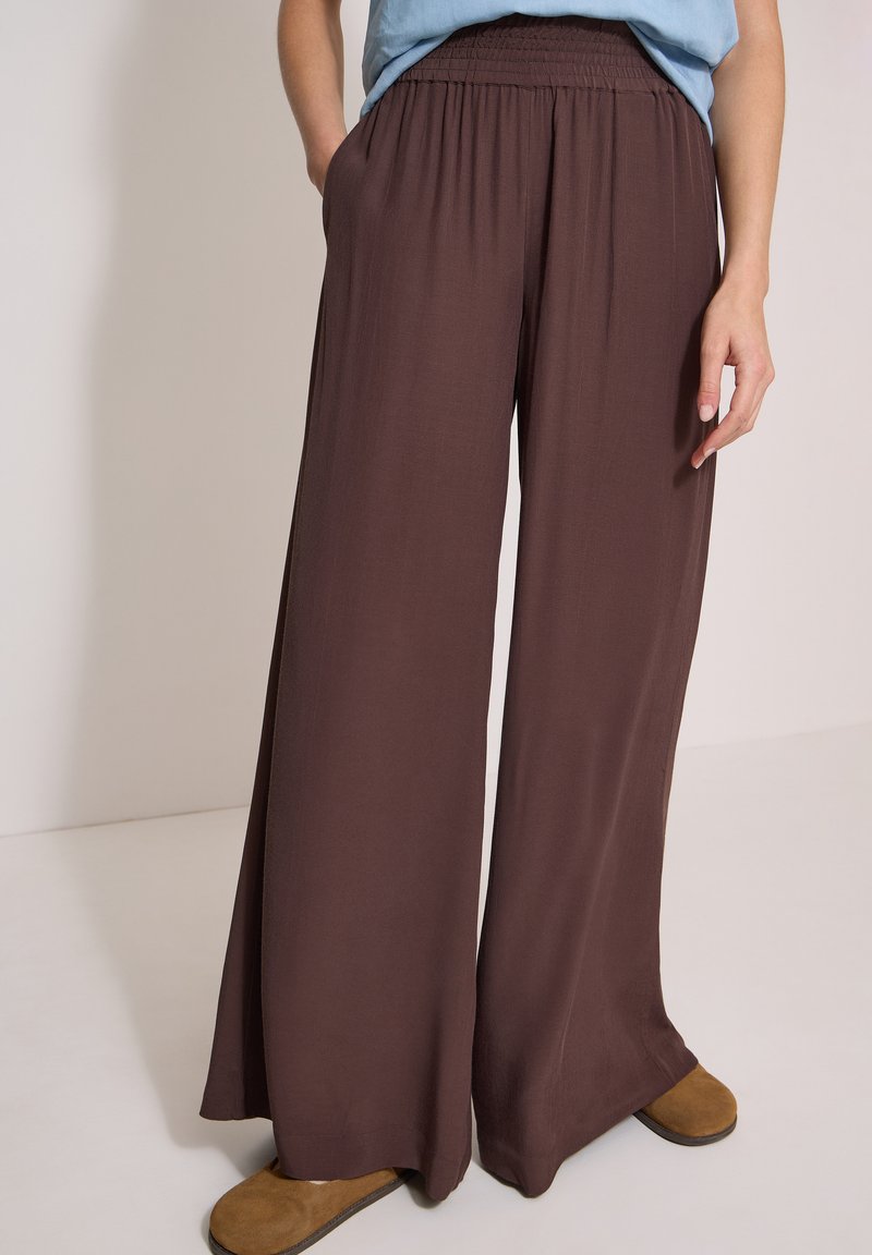 Personne portant un pantalon large marron, un haut bleu clair rentré et des chaussures plates marron, debout avec une main dans la poche.