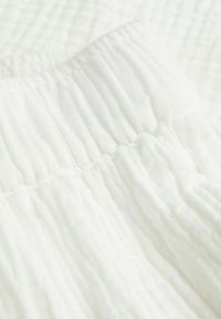 Bílý texturovaný materiál s pomačkaným vzorem a nařaseným elastickým pasem, který vykazuje jemný a vzdušný vzhled.