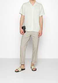 Camicia verde chiaro a maniche corte con bottoni, pantaloni beige a sigaretta e sandali a fascia neri e verde chiaro con dettagli rossi. Mano che tiene un portafoglio nero.