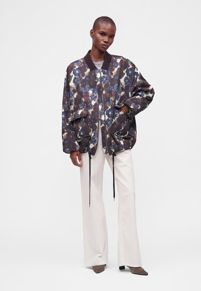 Personne portant une veste bomber oversize à fleurs foncées, un pantalon blanc à jambes larges, et des chaussures marron pointues, debout avec une main dans la poche de la veste.