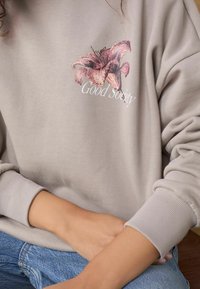 Hanorac gri cu un imprimeu floral texturat în nuanțe de roz și textul "Good Society" în design. Mansete cu nervuri și croială relaxată.