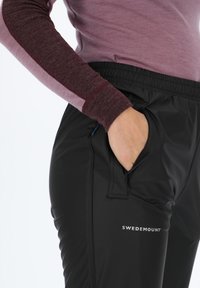 Pantaloni outdoor neri con design elegante e vita elastica. Presentano tasche con zip e un logo sulla parte inferiore della gamba. Il materiale appare liscio.