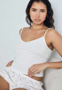 Witte geribde tanktop met dunne bandjes, voorzien van geschulpte randen, gecombineerd met witte shorts versierd met kleine rode hartjes.