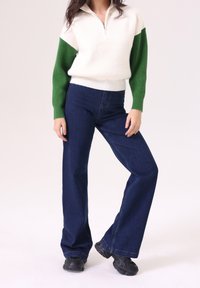 Suéter de punto blanco y verde con cuello de media cremallera, combinado con pantalones vaqueros de pierna ancha de color azul oscuro y calzado negro.