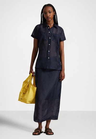 Sieviete ar gariem bizēm, valkājot tumši zilu īsroku pogājamu kreklu un atbilstošu garu svārku, tur dzeltenu auduma maisiņu, valkā brūnas sandales.