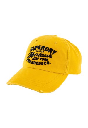 Casquette de baseball jaune avec texte brodé noir "Superdry Montauk New York The Goods Co." et petits trous usés sur la visière.