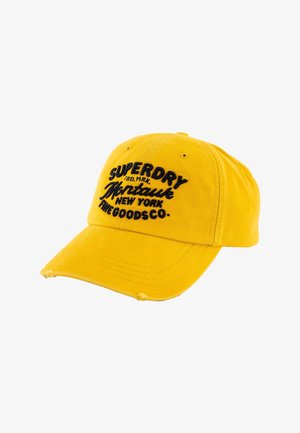 Gorra de béisbol amarilla con texto bordado en negro "Superdry Montauk New York The Goods Co." y pequeños agujeros desgastados en la visera.