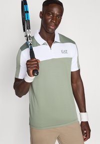 Πόλο πουκάμισο σε ανοιχτό πράσινο και λευκό με φερμουάρ στο γιακά· διαθέτει λογότυπο στο στήθος και υφή στην επιφάνεια. Κρατάει ρακέτα τένις.