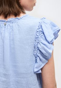 Γυναίκα με ανοιχτό μπλε μπλουζάκι με βολάν στα μανίκια και το γιακά, εμφανιζόμενη από πίσω και πλάγια σε ουδέτερο φόντο.