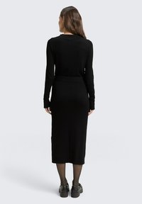 Langärmliges, schwarzes Kleid in voller Länge mit geripptem Textur, tailliertem Schnitt und Schlitzen am Rücken. Kombiniert mit schwarzen Schuhen und dünnen Strumpfhosen.