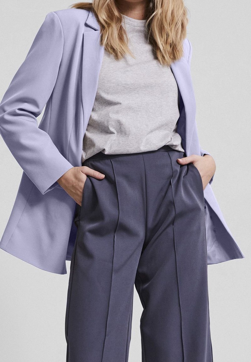 Žena v světle fialovém sakě, šedém tričku a tmavých kalhotách s vysokým pasem má ruce v kapsách před jednolitým pozadím.