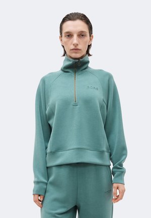 Young person wearing teal half-zip sweatshirt and matching pants with subtle "BORG" branding on chest and thigh.