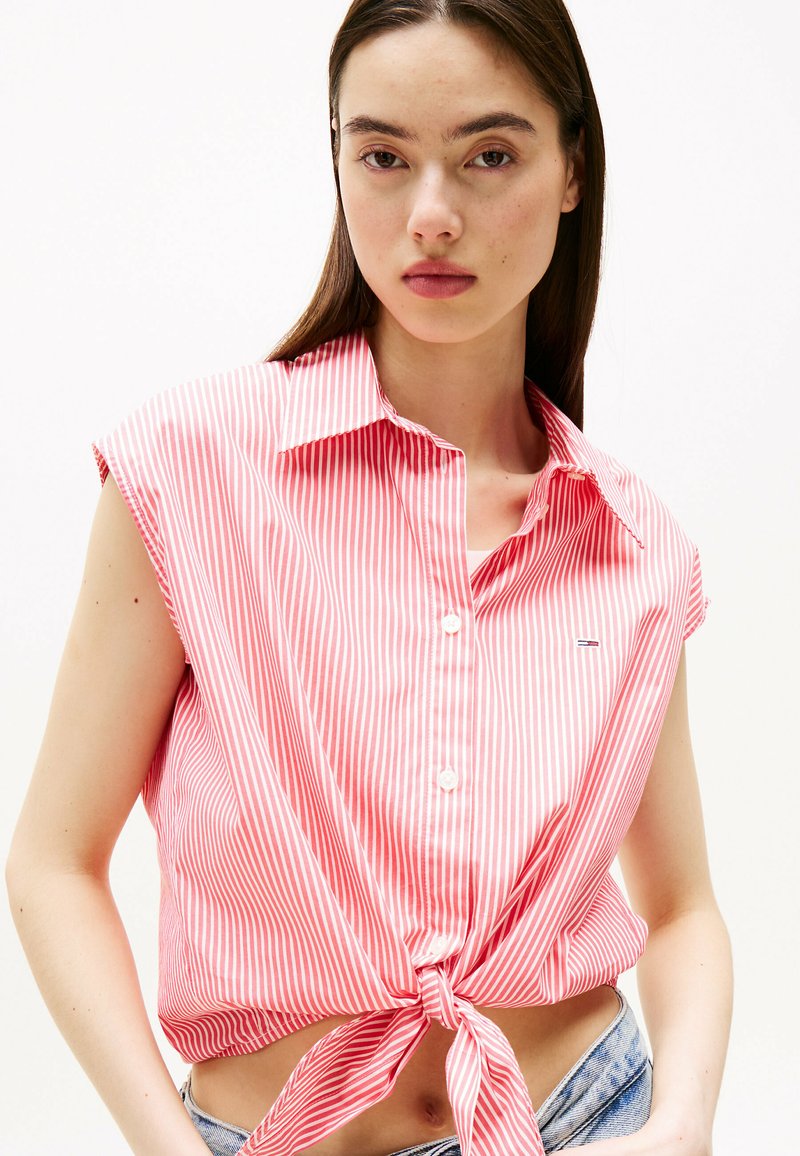 Chemise crop rayée rouge et blanche avec des manches courtes, un col, un boutonnage à l'avant et un nœud noué à la taille. Détail de petit logo sur la poitrine.