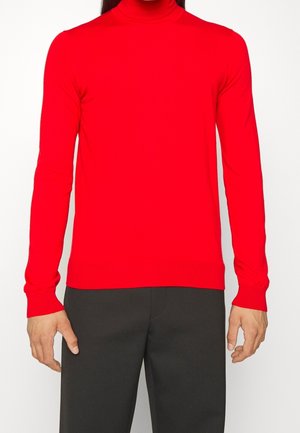 Red turtleneck sweater made of stretchy knit fabric. Features fitted long sleeves and ribbed cuffs and hem. Worn with black pants.