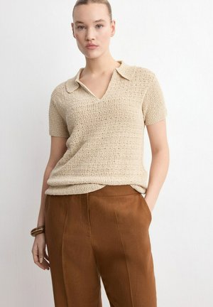 Femme portant un haut en maille beige à manches courtes avec col et un pantalon marron taille haute, debout devant un fond blanc uni.