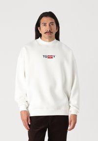 Bijela dukserica s visokim vratom, s "TOMMY" logotipom u mornarskoj plavoj i crvenoj boji na prsima. Mekana tekstura i opušteni rukavi.