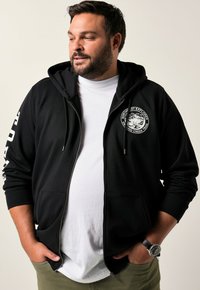 Sort zip-up hoodie med en frontlomme, logo p�å brystet og grafisk tekst ned ad ærmet; båret over en hvid t-shirt.