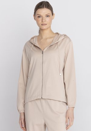 Kobieta w beżowej bluzie z kapturem zapinanej na zamek z troczkami i dopasowanych spodniach, stojąca na tle jednolitego jasnego tła.