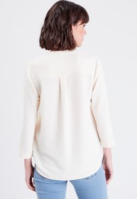 Blouse à manches longues en crème clair avec un dos texturé, présentant un pli au centre et un ourlet arrondi. Portée avec un jean bleu.