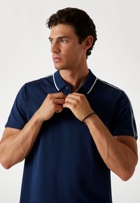 Marineblaues Poloshirt mit weißen Akzenten, Knopfleiste und kurzen Ärmeln. Der Stoff wirkt glatt; das Modell passt den Kragen mit beiden Händen an.