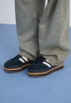 Personne portant un jean bleu clair ample et des baskets plateformes noires avec des rayures blanches, debout sur un tapis bleu.