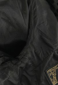 Tissu de veste matelassée noire avec coutures visibles et un petit patch rectangulaire présentant le contour stylisé d'un visage en fil doré.