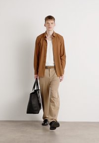 Camicia marrone a maniche lunghe sopra una canotta bianca, pantaloni khaki, borsa nera e scarpe nere, su uno sfondo bianco semplice.