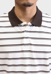 La polo presenta un tessuto bianco con righe orizzontali marroni, un colletto marrone scuro e due bottoni al collo. Design morbido e informale.