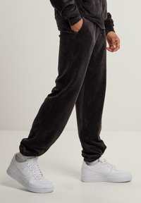 Melnas velūra džinsu bikses ar elastīgām aprocēm un sānu kabatām, apvienotas ar baltām Nike Air Force 1 kedām. Gluda faktūra un brīva piegriezuma stils.