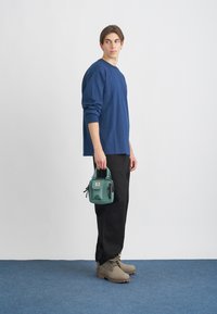 Sininen pitkähihainen paita, mustat housut, harmaat saappaat, pitää pientä vihreää laukkua. Yksivärisiä, rentoa muotoilua, yksinkertaisia muotoja ja tekstuureja.