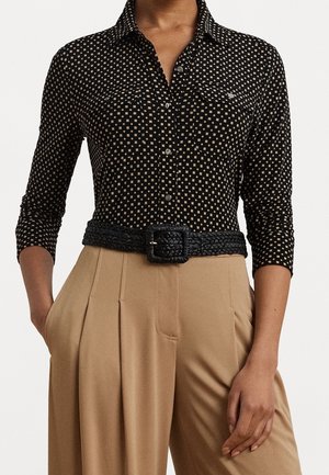 Vrouw draagt een zwart shirt met lange mouwen met kleine beige vierkantjes, gecombineerd met beige hoge taillebroeken en een brede zwarte gevlochten riem.