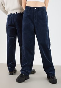 Tamno plavi kord pantaloni s opuštenim krojem, visokoga struka, prednjim džepovima i pohabanim detaljima na rubu straga, u kombinaciji s masivnim crnim cipelama.