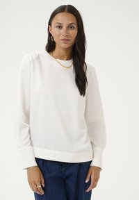 Witte blouse met lange mouwen en een ronde halslijn, gladde structuur en een lichte glans. Gecombineerd met donkerblauwe spijkerbroek. Accentuatie met een gouden ketting.