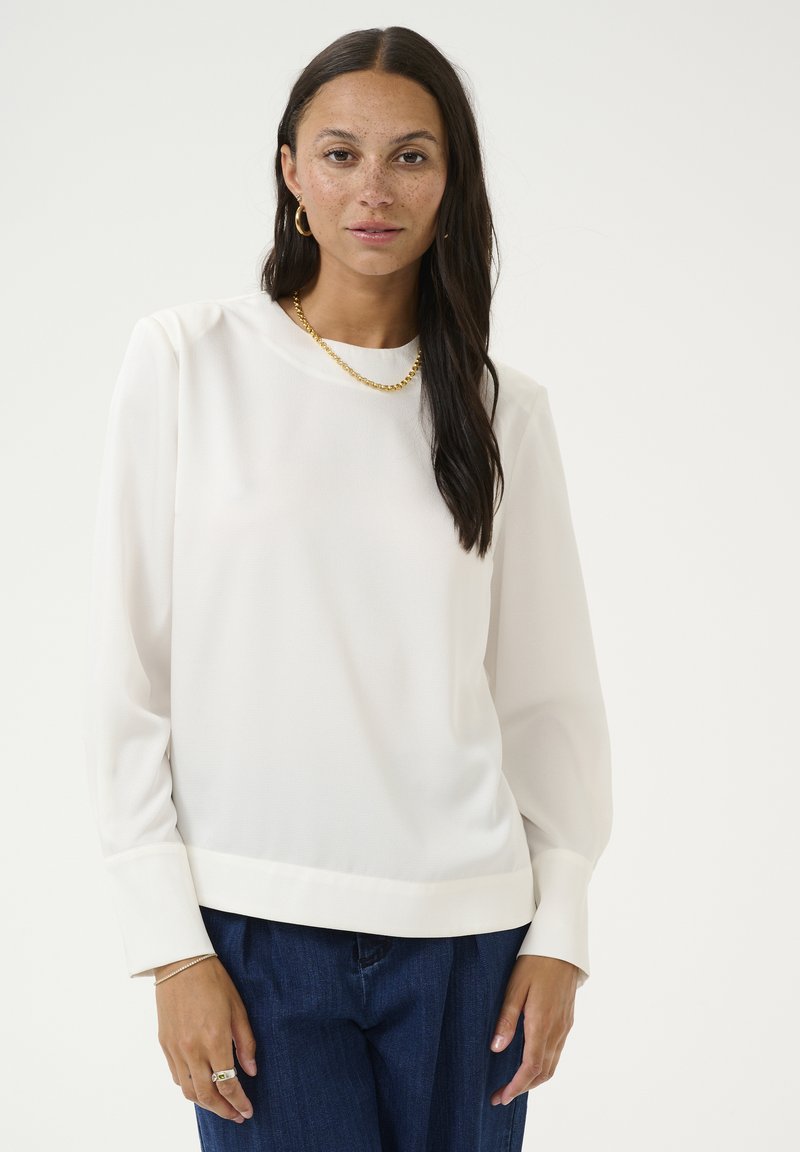 Witte blouse met lange mouwen en een ronde halslijn, gladde structuur en een lichte glans. Gecombineerd met donkerblauwe spijkerbroek. Accentuatie met een gouden ketting.