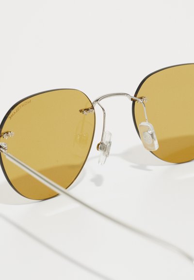 Lunettes de soleil teintées en jaune avec de grandes lentilles géométriques. Monture en métal argenté avec des plaquettes de nez et de petits accents décoratifs près des charnières.