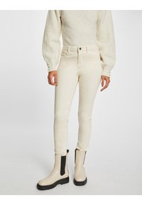 Jeans skinny beige de cintura alta, con bolsillos delanteros y traseros, combinados con botas de tobillo crema que tienen paneles laterales negros.
