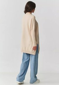 Beżowy oversize sweter z prążkowanymi mankietami i dołem, z bocznymi rozcięciami, zestawiony z jasnoniebieskimi spodniami z szerokimi nogawkami i białymi butami.