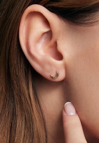 Złoty kolczyk w kształcie półksiężyca, o gładkiej powierzchni, mały rozmiar, na tle zbliżenia ucha z brązowymi włosami i ręką delikatnie dotykającą skóry.