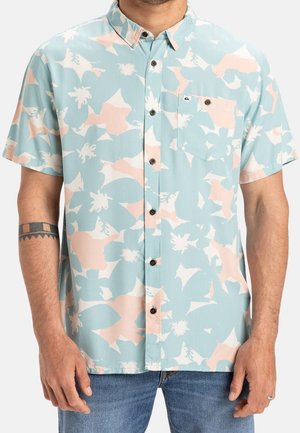 Man draagt een kortärmelig overhemd met grote abstracte bloemenprint in lichtblauw, lichtperzik en wit, gecombineerd met een blauwe spijkerbroek.