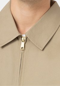 Beżowa kurtka z kołnierzykiem i złotym zamkiem błyskawicznym oraz fakturowaną tkaniną, charakteryzująca się czystym designem i ostrymi krawędziami, podkreślającymi solidne wykonanie.