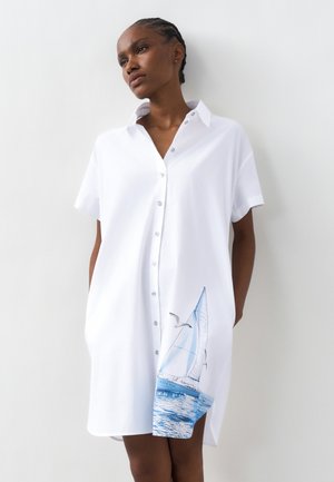 Kvinde med fletninger, iført en hvid kjole med knapper og et motiv af en blå sejlbåd og en måge nær kanten, stående foran en hvid væg.