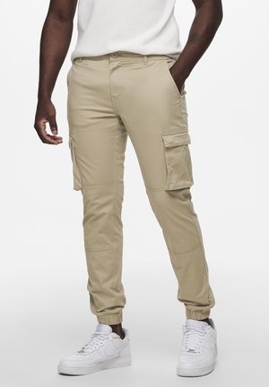 Beige cargobroeken met zijzakken en elastische boorden, gemaakt van lichte stof. Gepaard met witte sneakers voor een casual uitstraling.