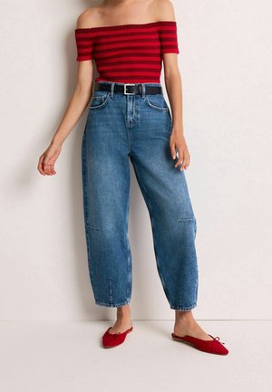 Vrouw draagt een rood en zwart gestreept off-shoulder top, hooggetailleerde losse blauwe jeans met zwarte riem en rode platte instapschoenen.