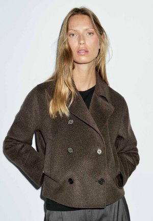 Young woman with long blonde hair wears a double-breasted dark brown coat over a black top, standing against a plain white background.
