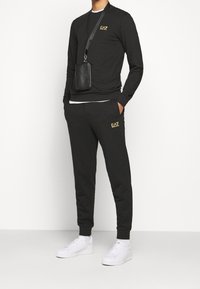 Svart långärmad tröja med guldlogotyp, matchande svarta joggingbyxor och vita sneakers. Modellen bär en liten svart väska med rem.