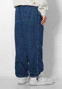 Blå denim cargo byxor med avslappnad passform, elastiska muddar, sidofickor och en texturerad tyg. Parat med vita sneakers.