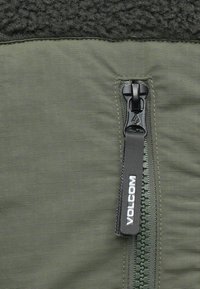 Green hybrid jacket featuring a smooth, flexible fabric with a fuzzy black fleece upper section and a black zip closure with a logo tag.